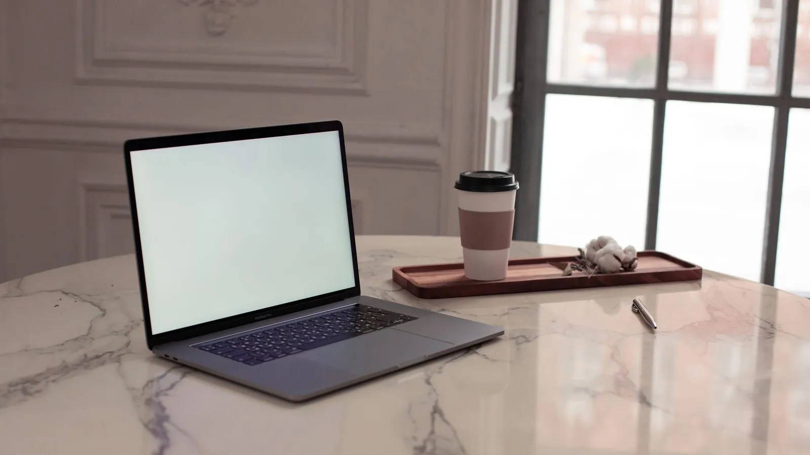 A laptop and a takeaway coffee on a bright marble table beside a tall window