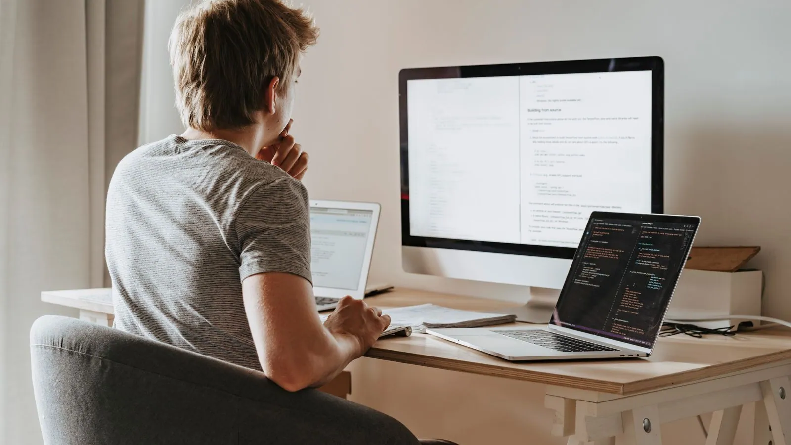 A developer at a bright desk with a large monitor and a laptop, mid-build